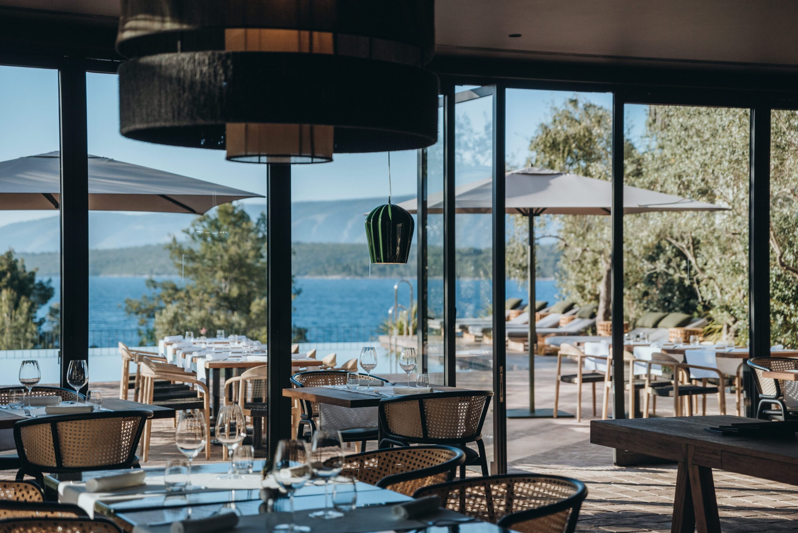 MaslinaResort_TheRestaurant_09_KateSevo Elegant, minimalist-style restaurant with floor-to-ceiling windows and a poolside terrace, offering panoramic views of the Adriatic Sea; features natural materials like terracotta and wood, reflecting the surrounding olive groves and vineyards.